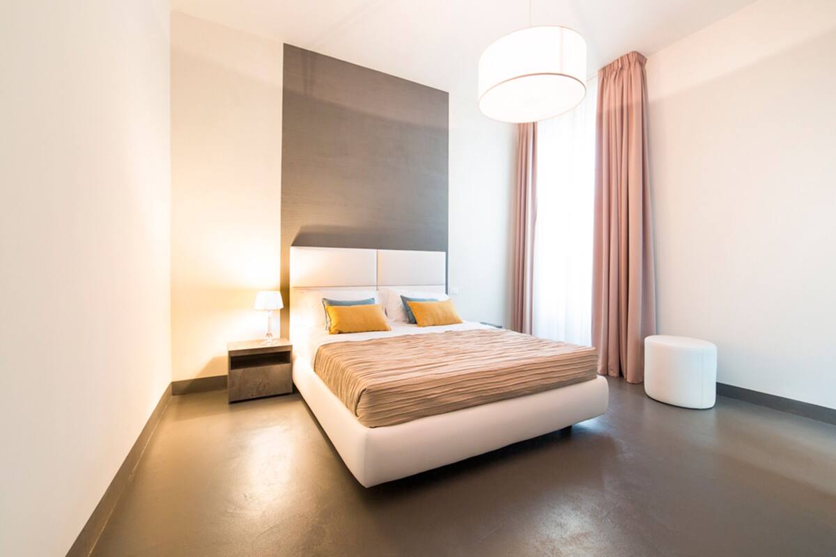 Una habitación de hotel moderna con una cama grande, una mesita de noche, una lámpara y un amplio ventanal con cortinas.