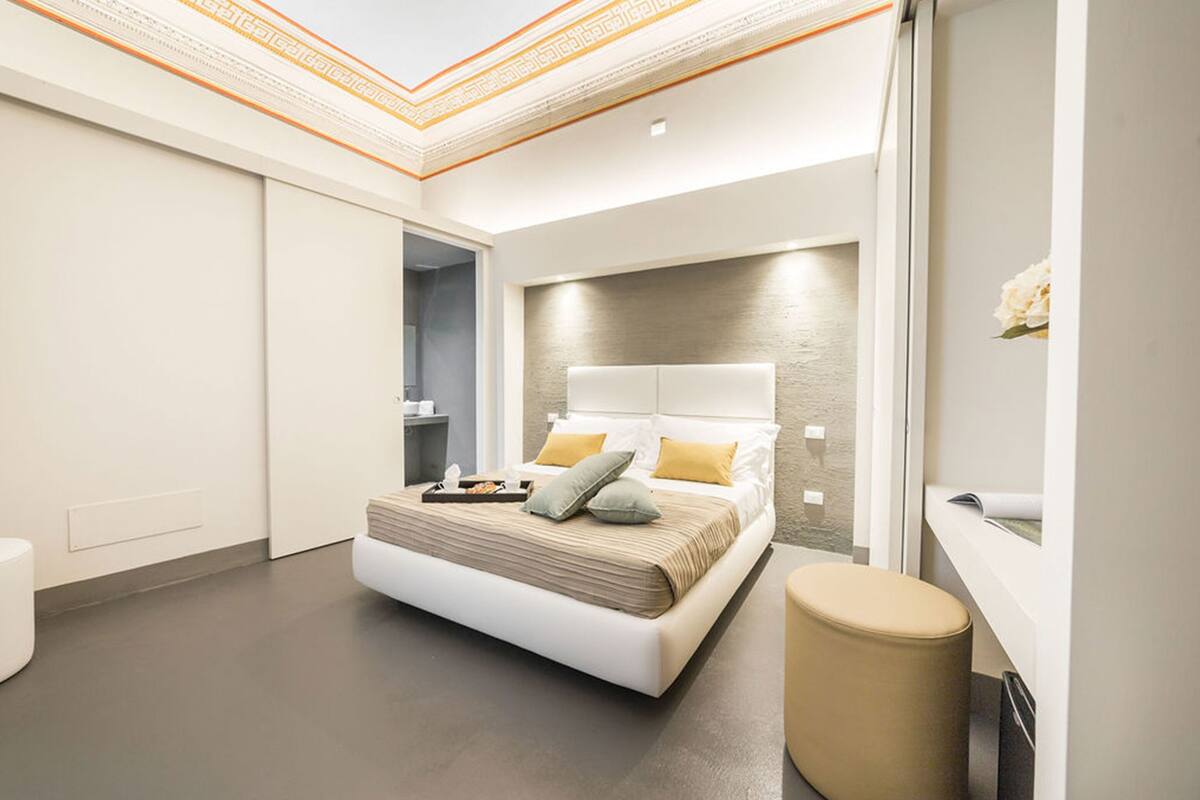 Un dormitorio moderno con una cama grande, una mesita de noche, un taburete pequeño y un ventanal con vistas.