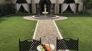 A dining table with a three-tiered fruit and pastry display, a fountain in the background, and a well-maintained lawn. - LUCUS Exclusive B&B (Lucca)