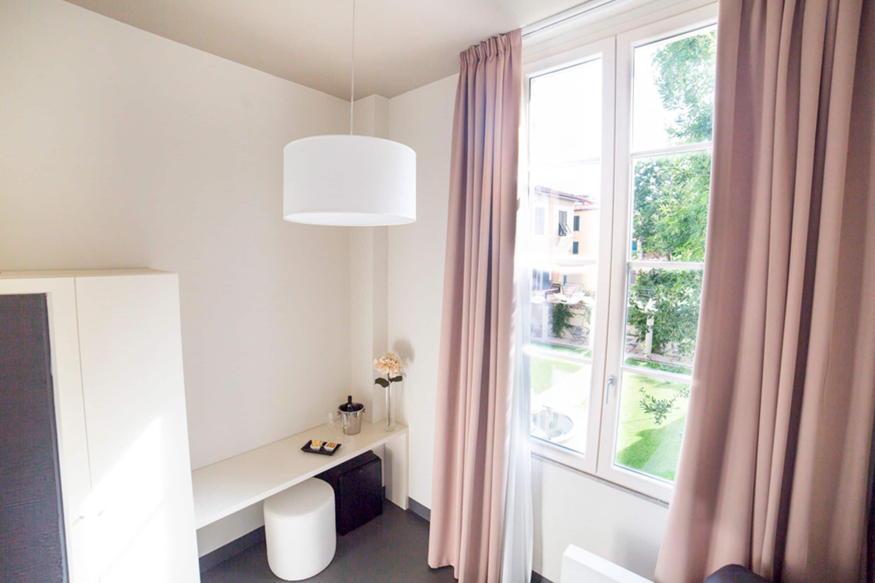 Een moderne hotelkamer met een groot raam, een wit bureau, een klein bankje en een hanglamp.