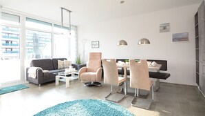 Ein modernes Wohnzimmer mit einer Couch, Sesseln, einem Couchtisch und einem Essbereich mit Tisch und Stühlen. Zwei Pendelleuchten befinden sich über dem Esstisch, und ein großes Fenster bietet Blick auf Gebäude.