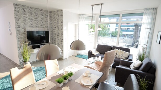 Ein modernes Wohnzimmer mit einem Flachbildfernseher, bequemen Sitzmöglichkeiten, einem Essbereich mit Tisch für zwei Personen und großen Fenstern mit Stadtblick.