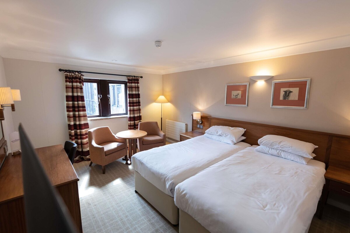A hotel room with two beds, a desk, a chair, a small table, and a window with curtains.