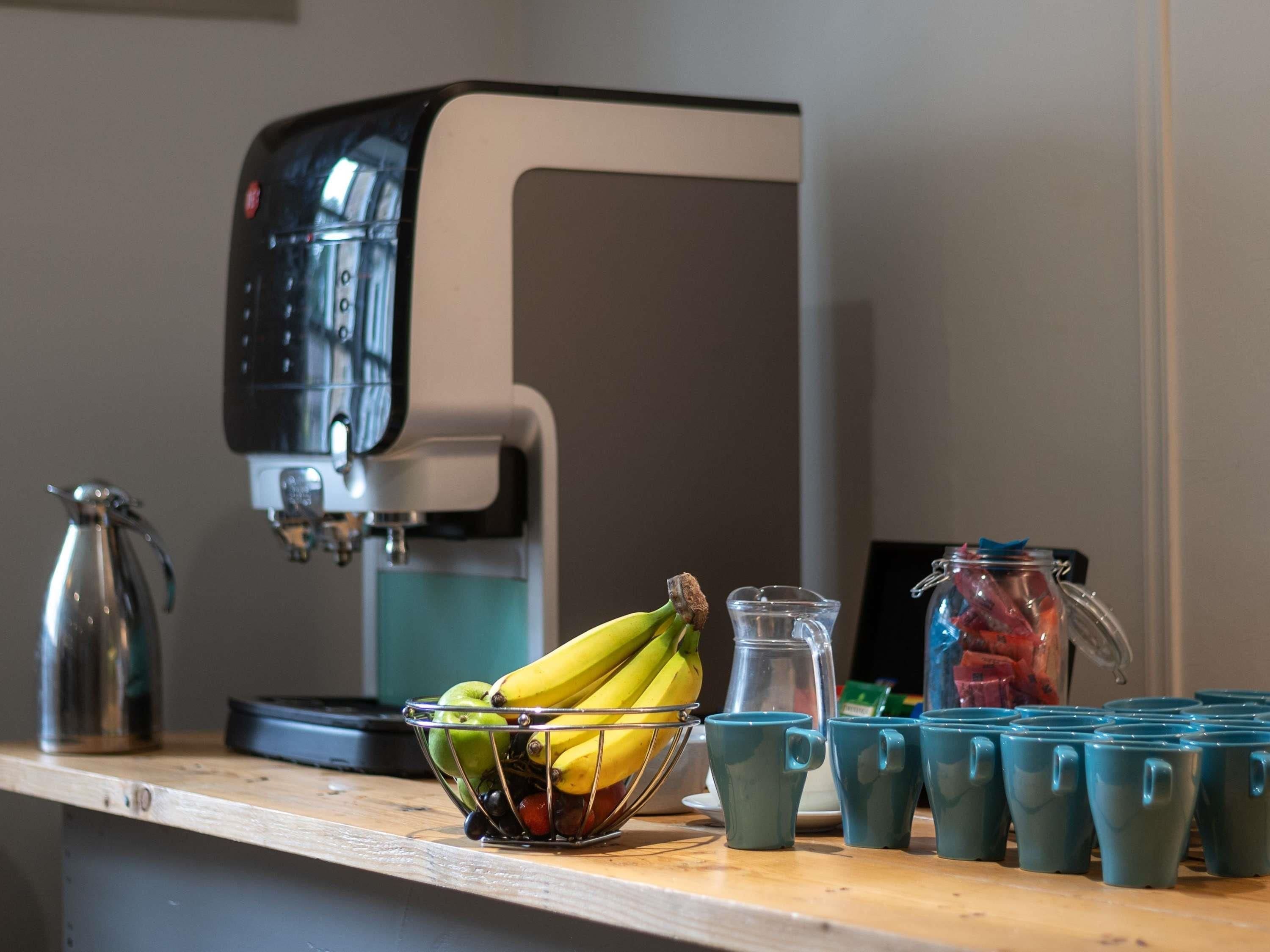 Une machine à café, un panier de fruits contenant des bananes et des pommes, et plusieurs tasses de couleur turquoise sur un comptoir.