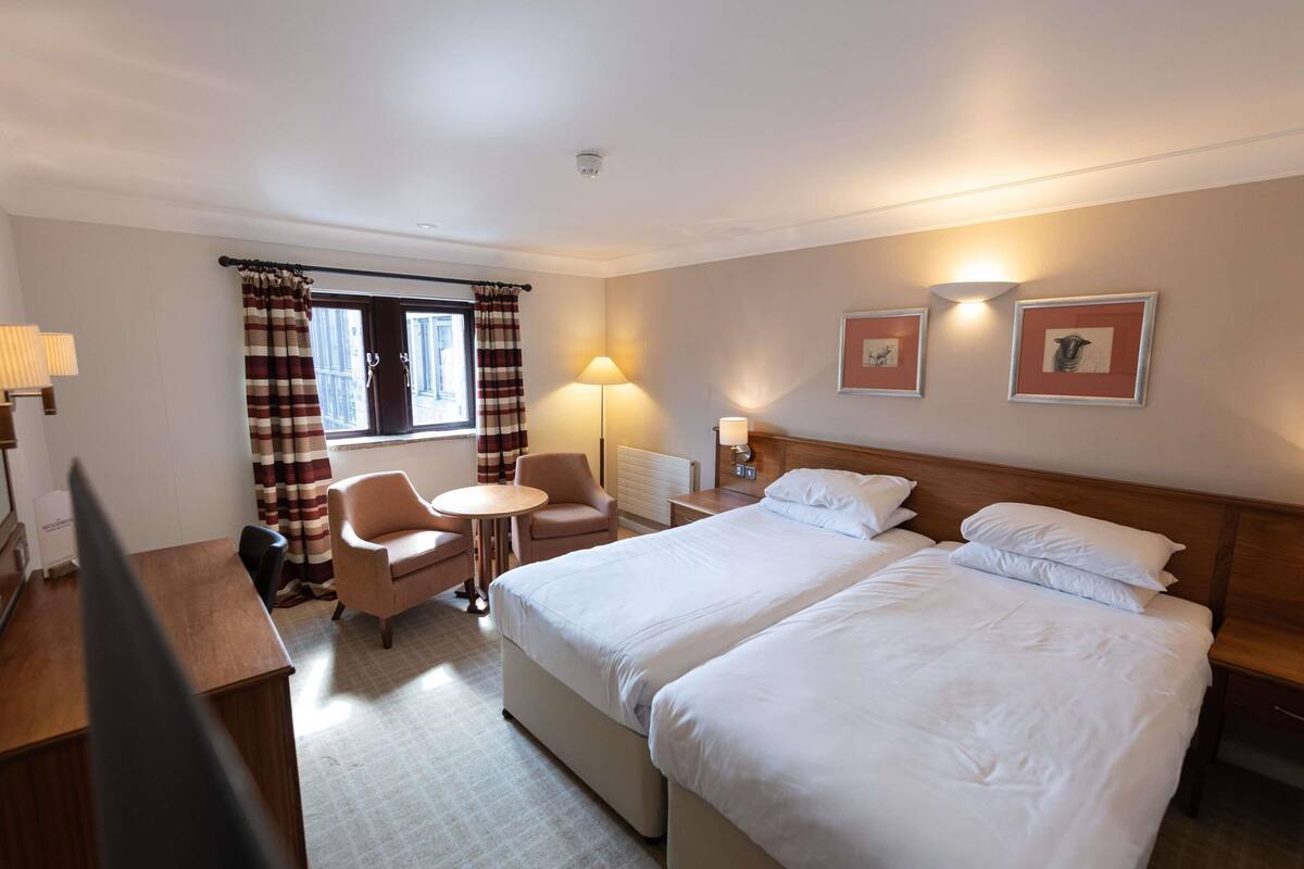 Habitación de hotel con dos camas, un escritorio, una silla, una mesita y una ventana con cortinas.