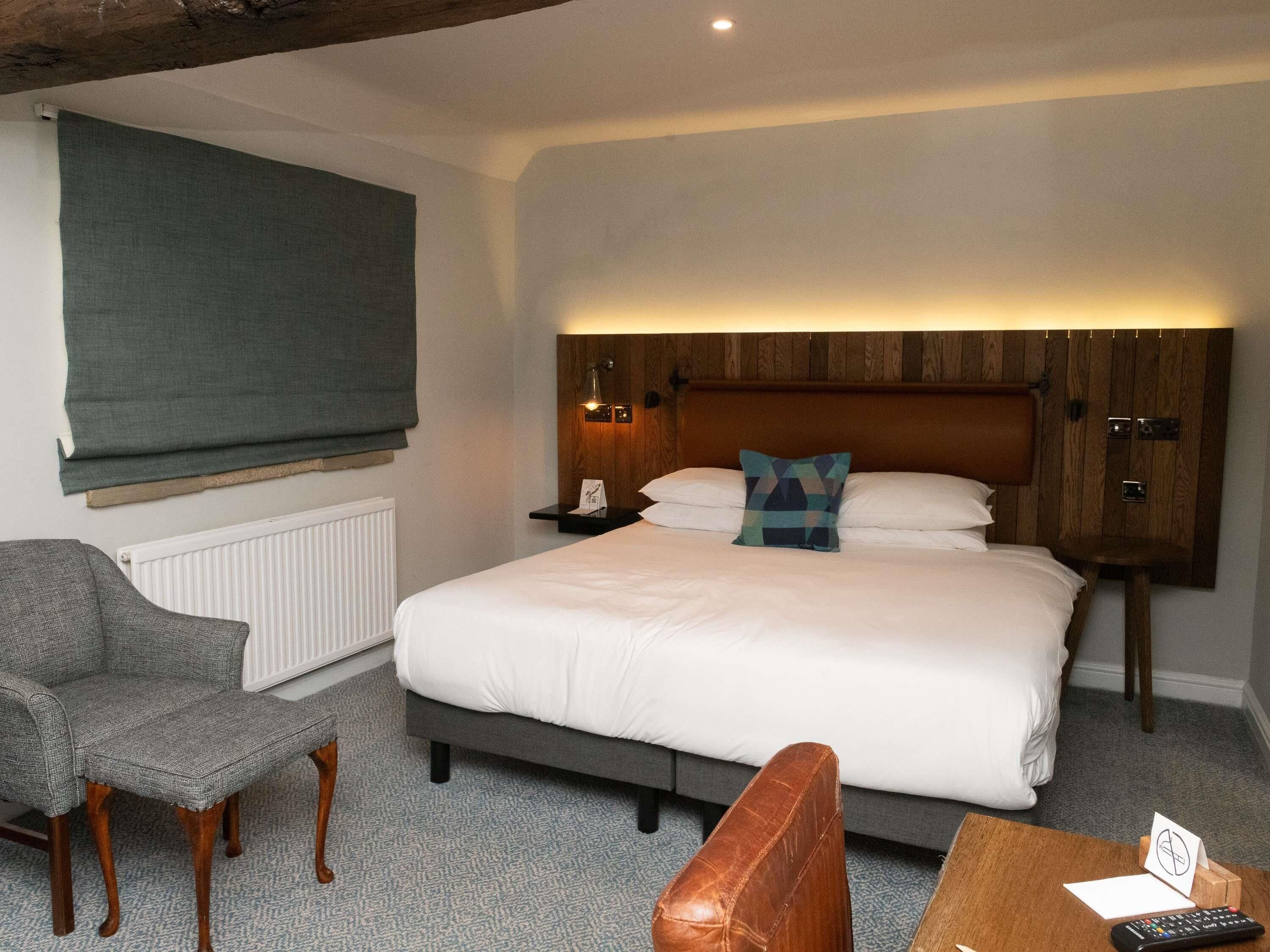 A hotel room with a bed, a chair, a small table, a nightstand, and a wall-mounted shelf.