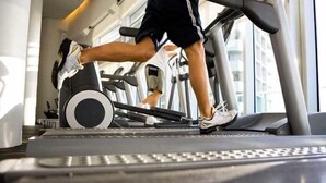 Uma pessoa corre numa esteira numa academia, com equipamentos de exercício e janelas ao fundo.