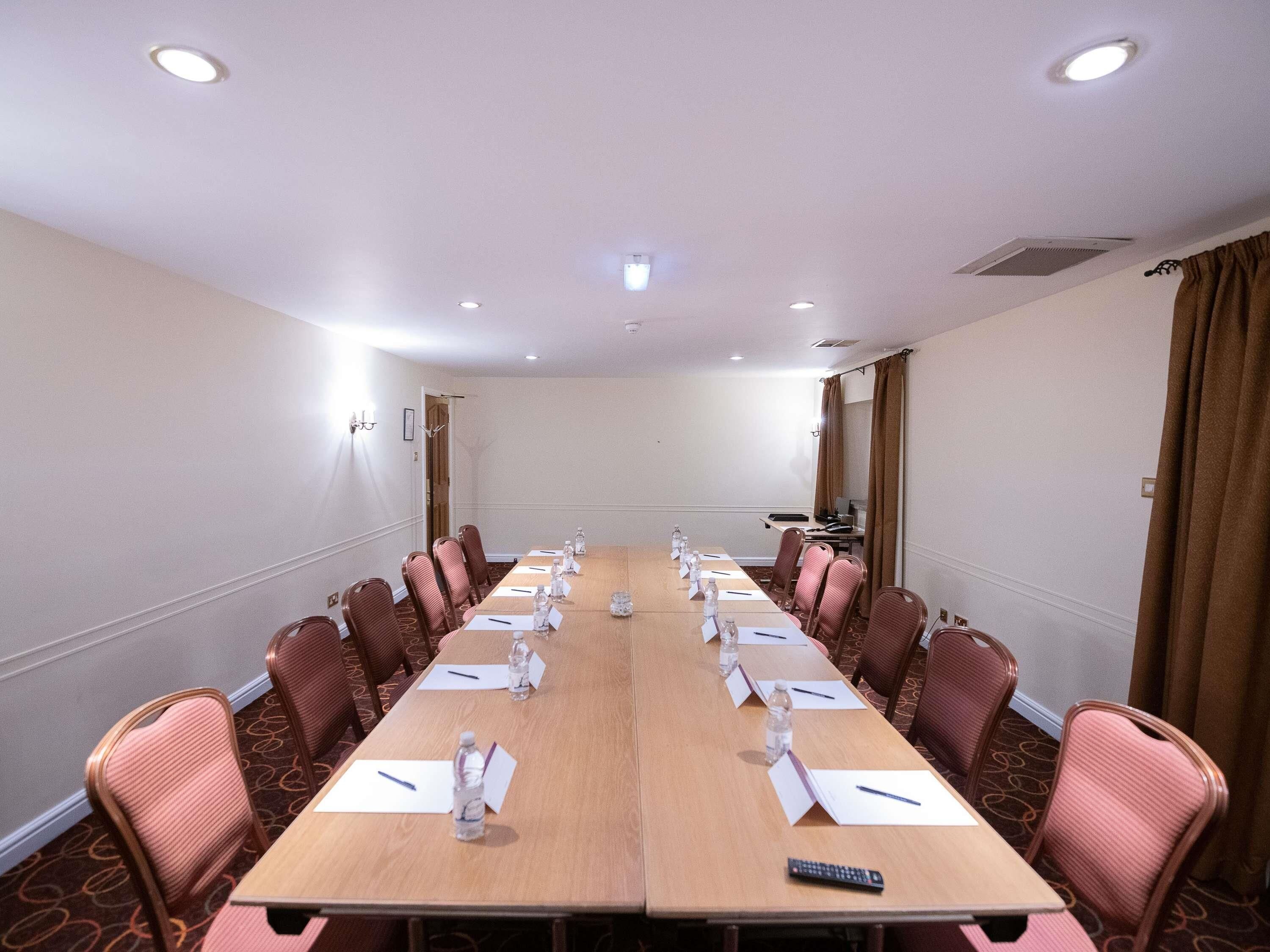 Una sala de conferències amb una taula llarga, cadirees, ampolles d'aigua, matisos de noms i un comandament a distància.
