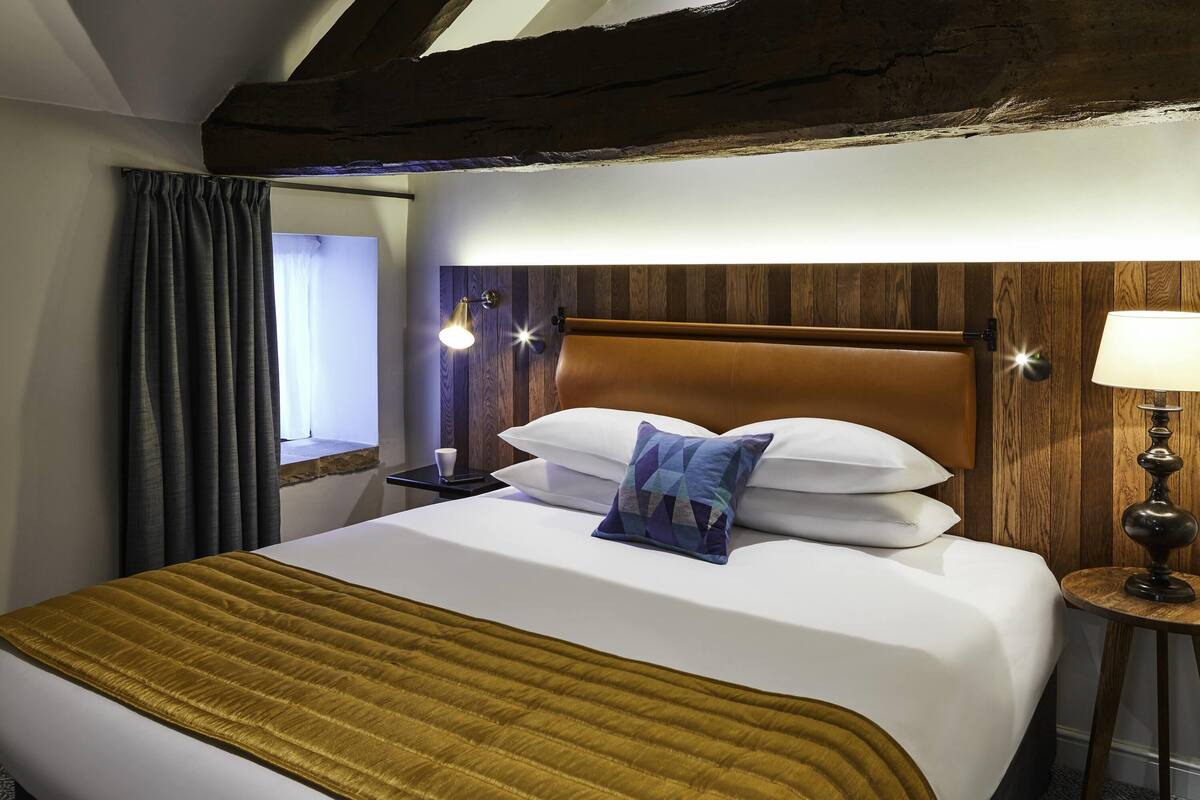 Habitación de hotel con una cama grande, cabecero de madera, una mesita de noche con lámpara y ventana con cortinas.
