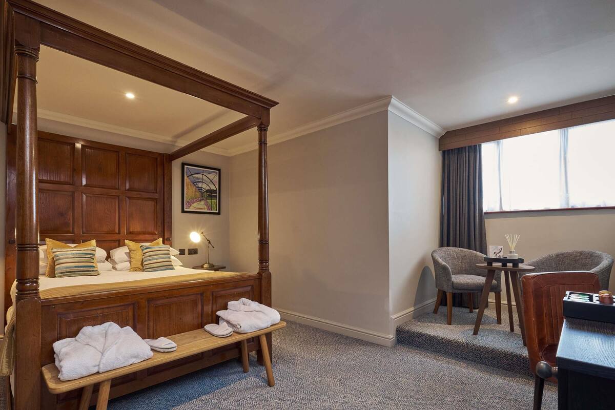 Una cama con dosel, un banco, un escritorio con sillas y una ventana con cortinas.