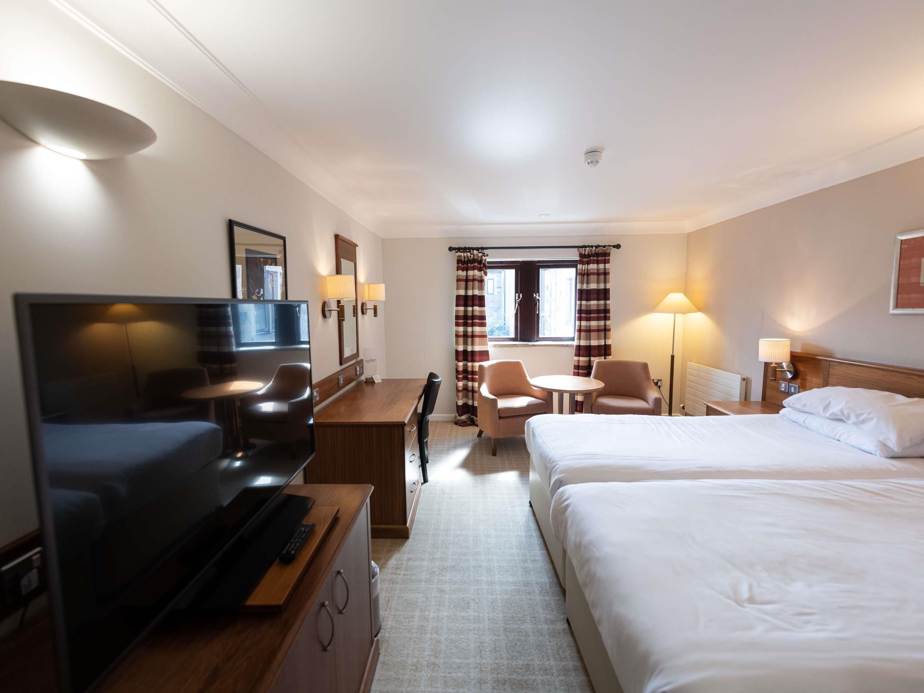 A hotel room with a TV, desk, two chairs, a small table, a bed, and a window with curtains.