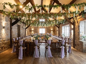 A rustic hall with stone walls, wooden beams, and a long table set for a formal event.