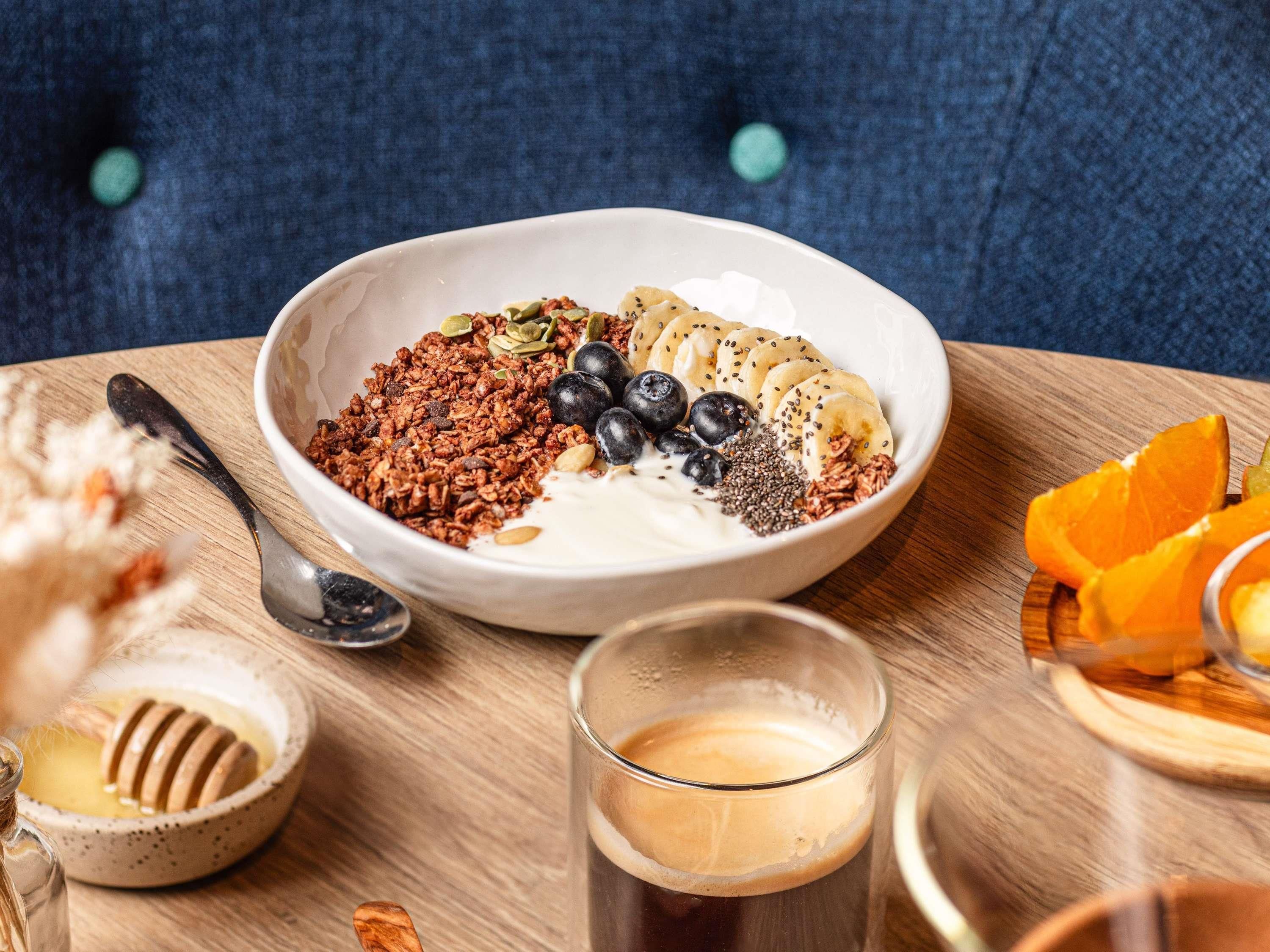 Un tazón de yogur con granola y fruta, una taza de café y un gotero de miel sobre una mesa de madera.