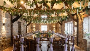 A rustic hall with stone walls, wooden beams, and a long table set for a formal event.