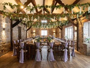 Un salón rústico con paredes de piedra, vigas de madera y una larga mesa dispuesta para un evento formal.