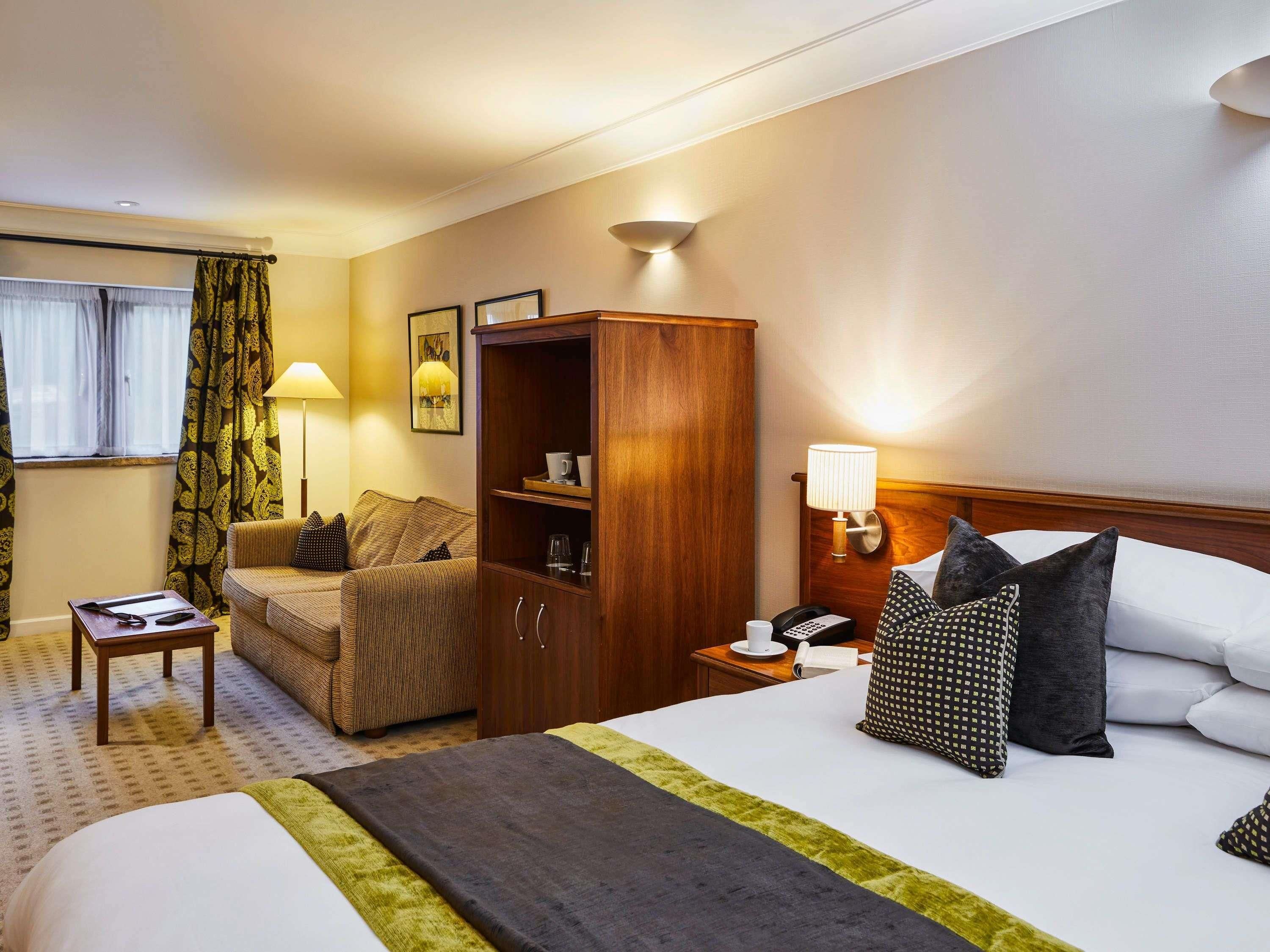 A hotel room with a bed, a sofa, a wooden cabinet, a nightstand, a lamp, a telephone, and a window with curtains.
