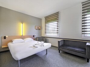 A hotel room with a bed, a sofa, a wall-mounted light, and a window with blinds. - ibis budget Katowice Centrum (Katowice)