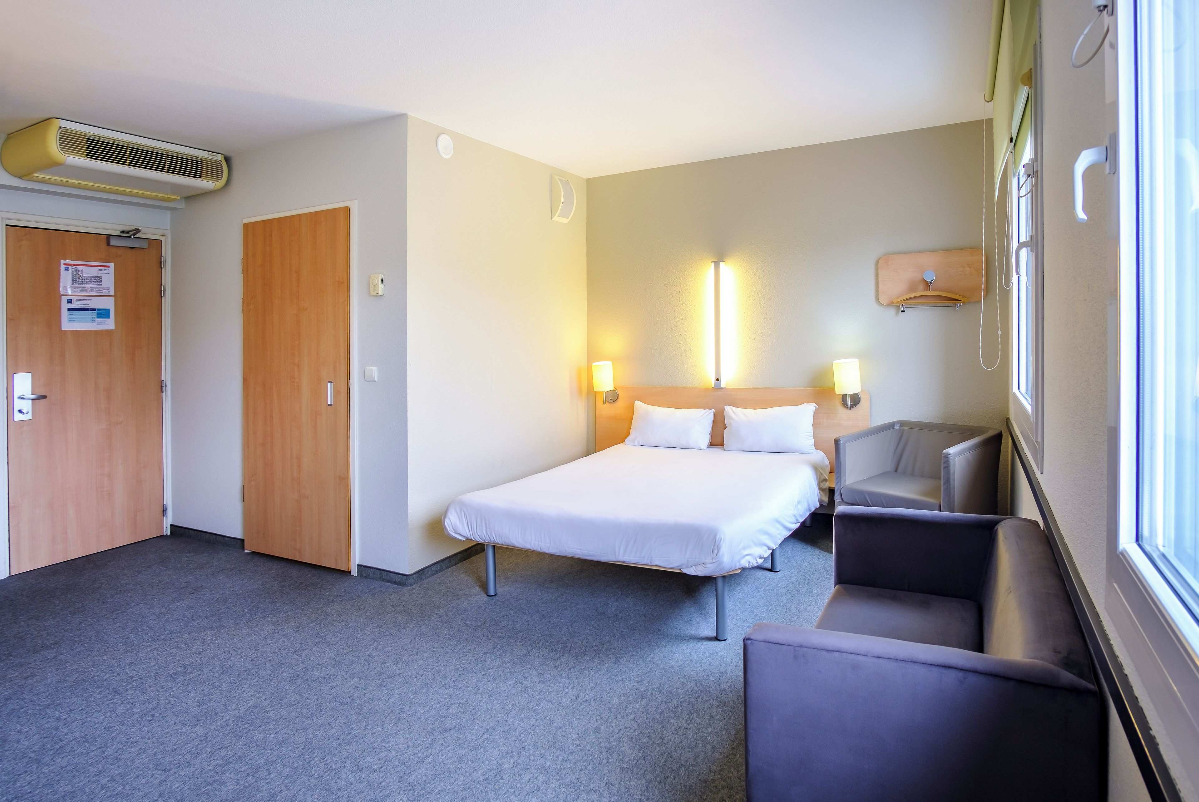 A hotel room with a bed, a desk, a chair, a window, and a door.