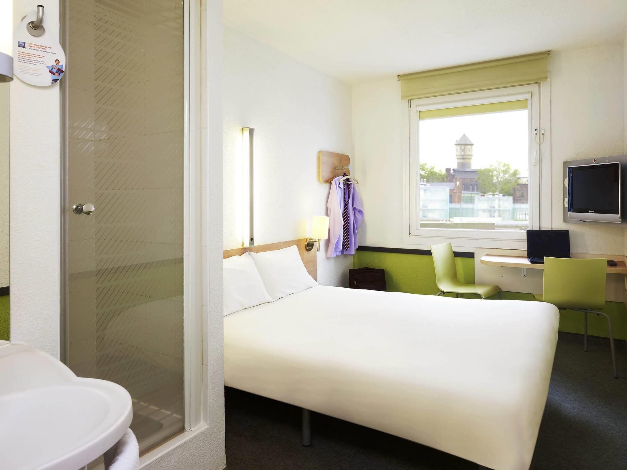 A hotel room with a bed, a desk with a computer, a TV, and a window with a view of buildings.