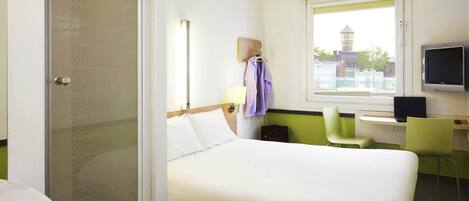 Habitación de hotel con cama, escritorio con computadora, televisor y ventana con vista a edificios.