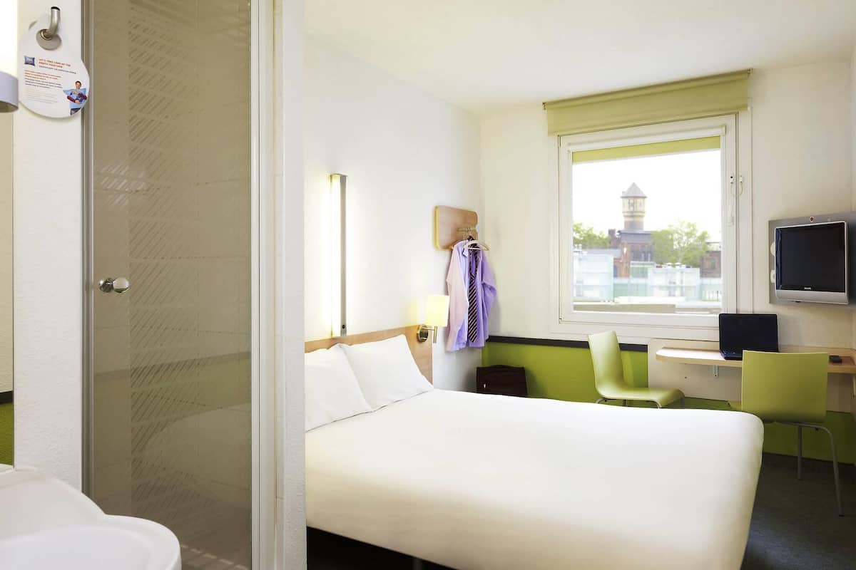 A hotel room with a bed, a desk with a computer, a TV, and a window with a view of buildings.