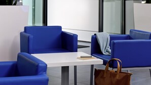 Un área de espera moderna con sillas azules, una mesa blanca y un bolso marrón en el suelo.