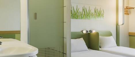 Habitación de hotel con cabina de ducha de vidrio, lavamanos con toalla, cama con ropa blanca y una lámpara fijada en la pared.