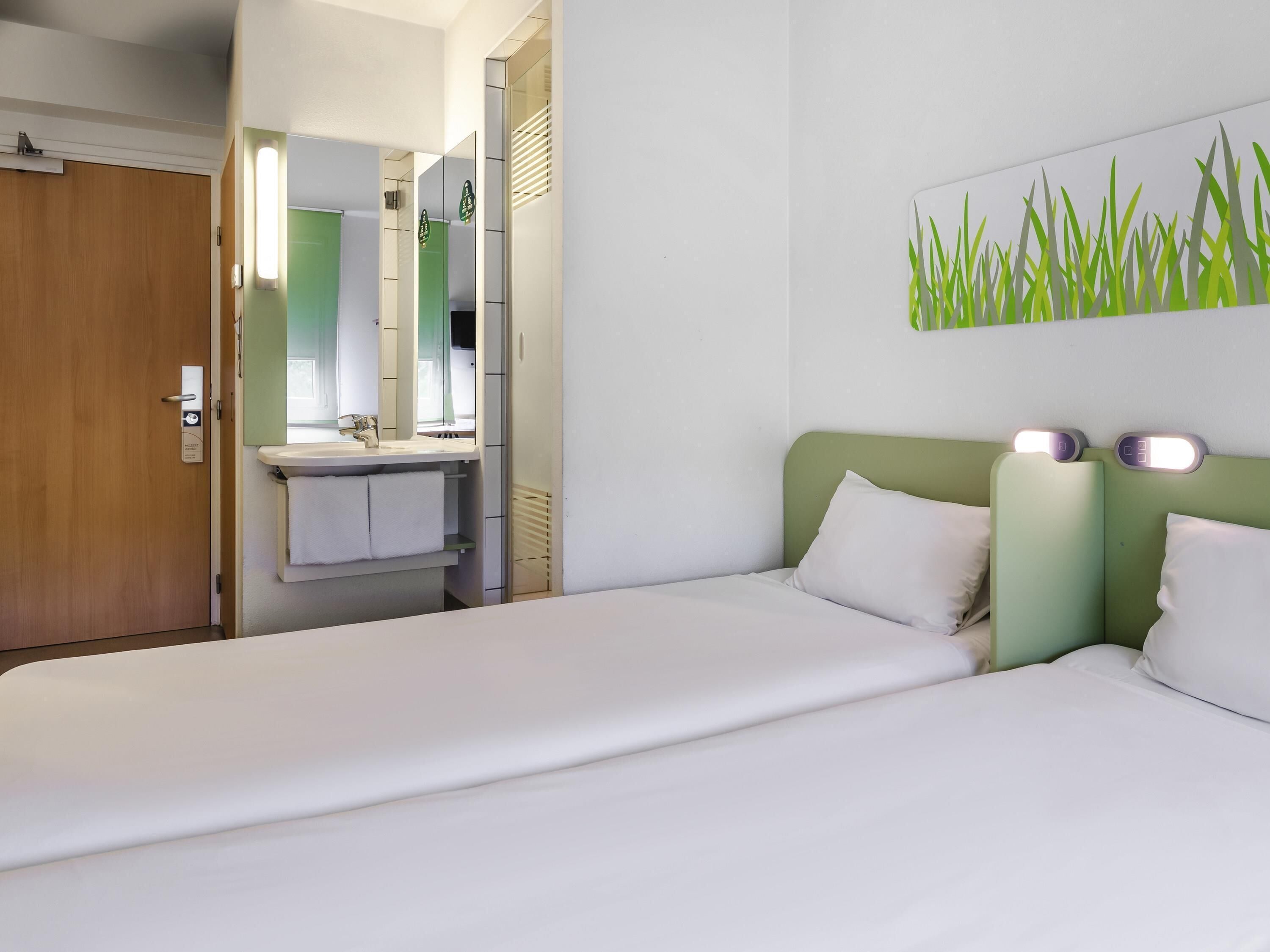 A hotel room with a bed, a bedside table, a mirror, a sink, and a green wall with a grass pattern.