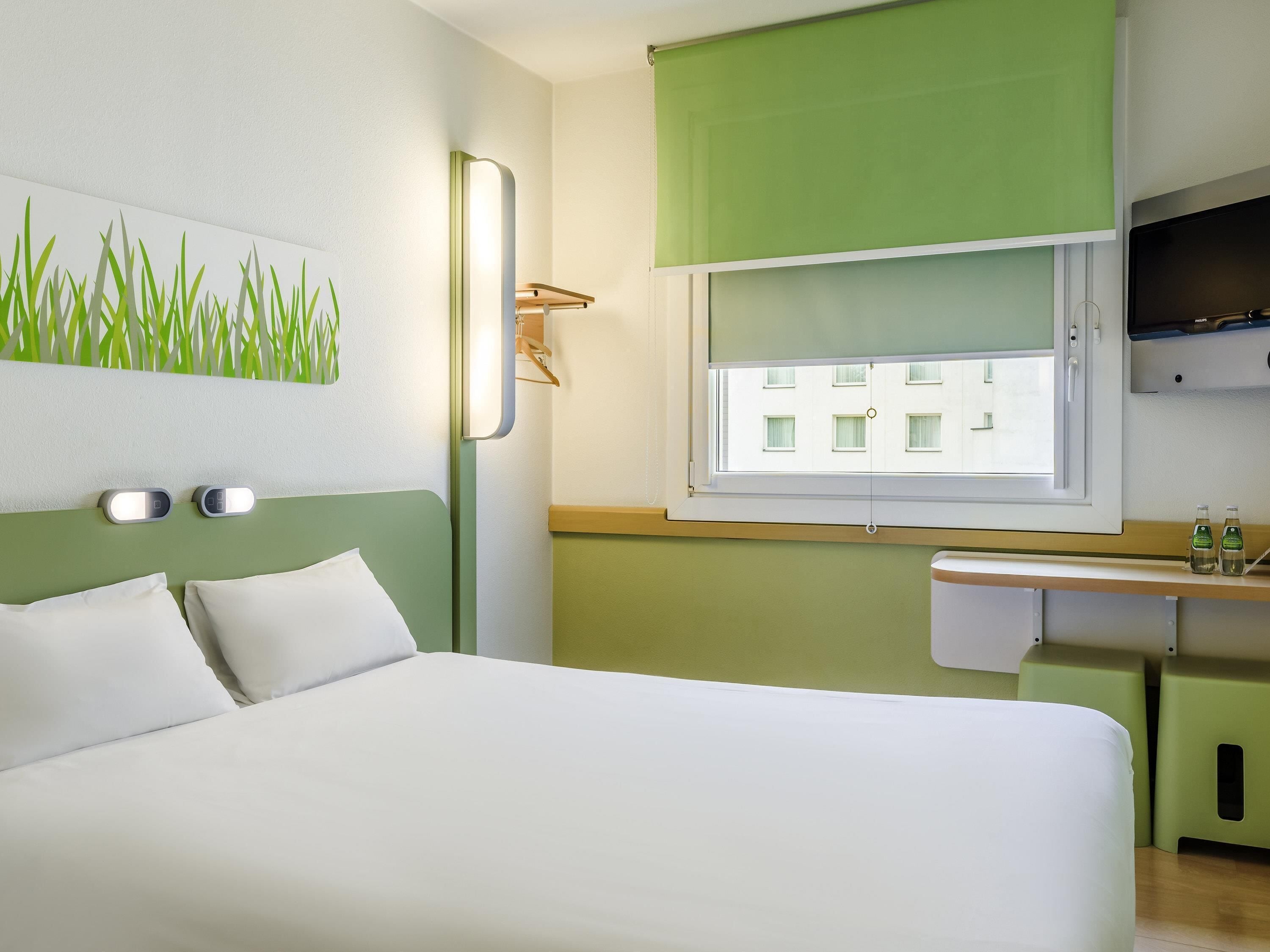 Une chambre d’hôtel avec un lit, une lampe fixée au mur, une fenêtre donnant sur des immeubles et une petite étagère avec des bouteilles.