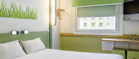 A hotel room with a bed, a wall-mounted light, a window with a view of buildings, and a small shelf with bottles.