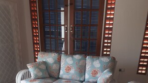 Un canapé en osier blanc recouvert d’un tissu à motifs floraux, une table basse en verre et une vue à travers une porte en bois dotée de panneaux de verre.