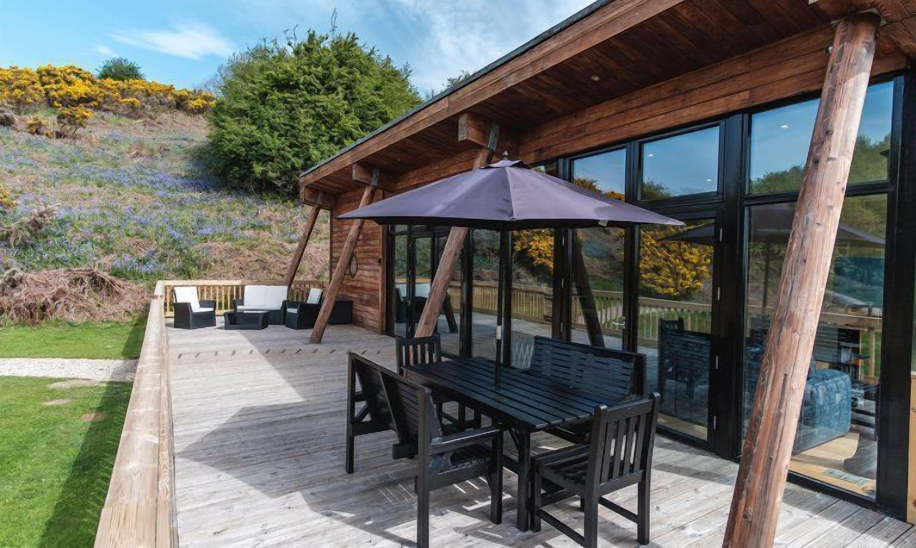 A wooden deck with a table and chairs, a covered seating area, and a glass enclosure with a view of the surrounding landscape.
