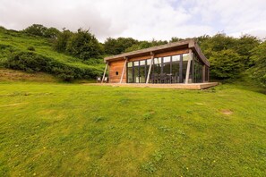 Een moderne houten cabin met grote ramen, gelegen op een grasachtige heuvel met een dicht bos op de achtergrond.
