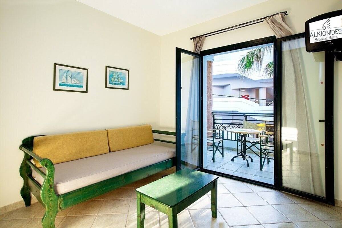 A room with a green sofa, a small table, and a view of the outside balcony with a table and chairs.