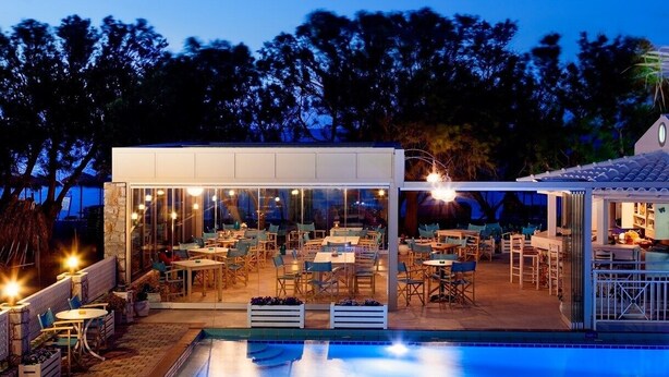 An outdoor dining area with tables and chairs, a pool, and a building with large windows. - Alkionides Seaside Apartments (Platanias)