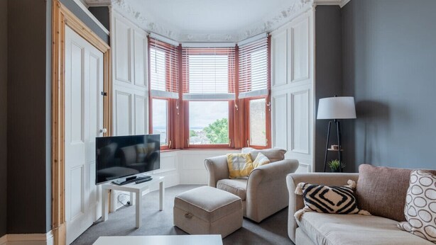 A modern living room with a television, two sofas, a coffee table, and a bay window with shutters.