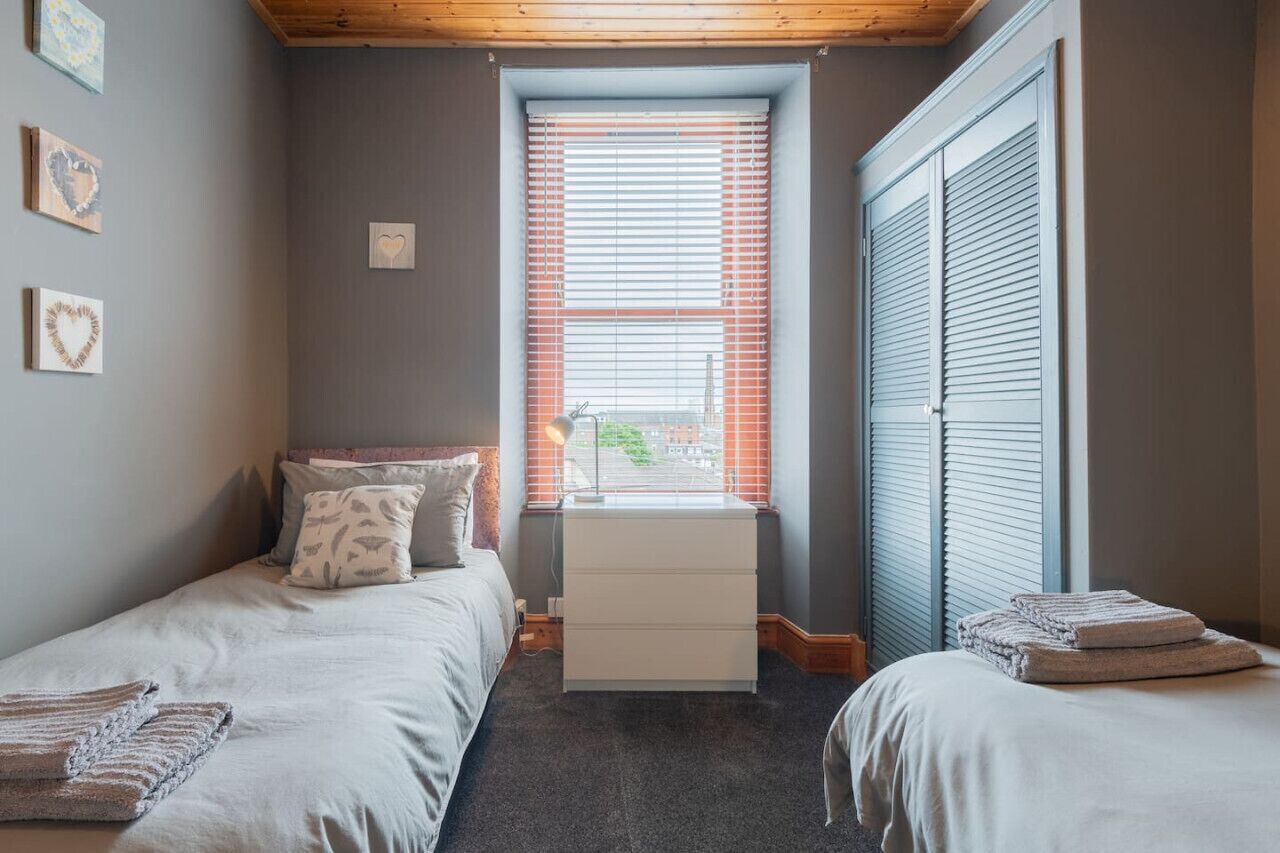 Ein Schlafzimmer mit zwei Einzelbetten, einer weißen Kommode, einem Fenster mit Jalousien und einem Kleiderschrank.