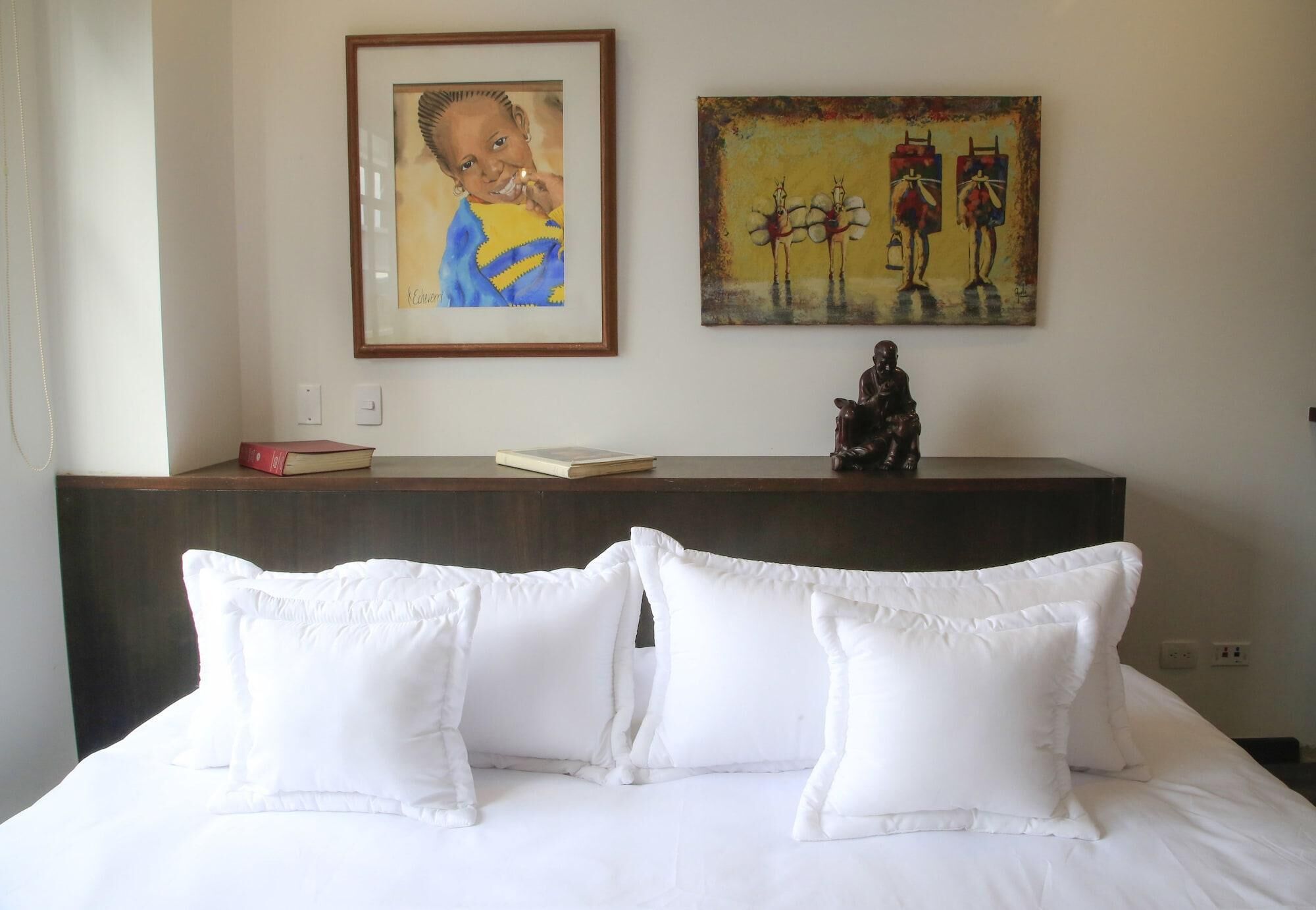 Habitación de hotel con cama, almohadas, mesita de noche con libros, un cuadro enmarcado y una estatua.