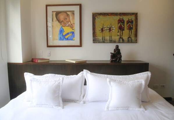 A hotel room with a bed, pillows, a nightstand with books, a framed painting, and a statue.