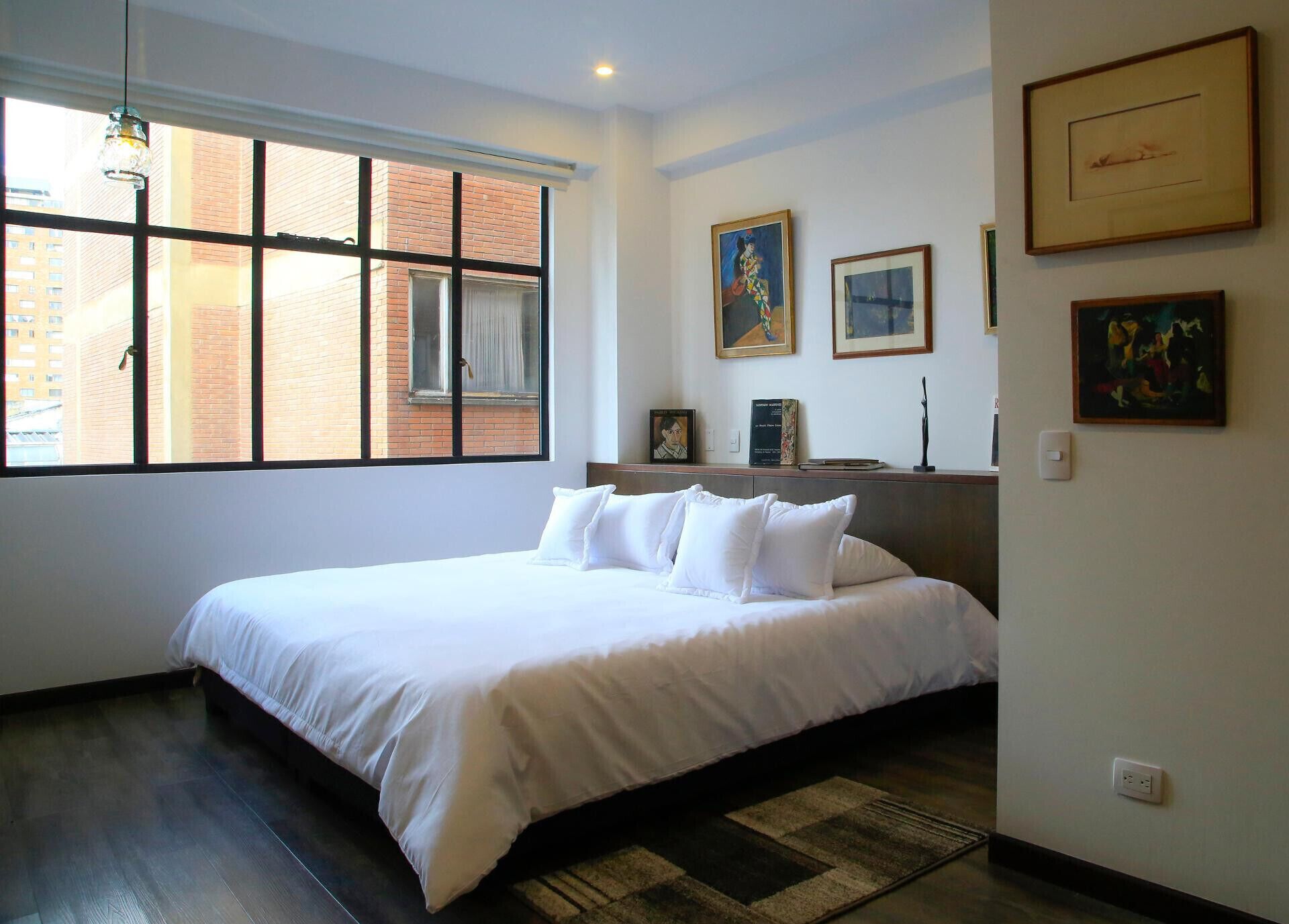 A hotel room with a large bed, a window with blinds, a wooden headboard, and several framed artworks on the wall.