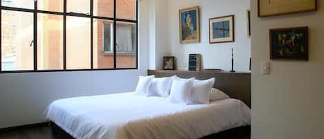 Habitación de hotel con una cama grande, una ventana con persianas, cabecera de madera y varias obras de arte enmarcadas en la pared.
