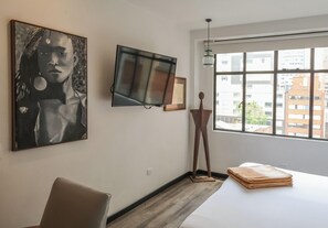 A room with a bed, a TV, a painting, and a window with a view of buildings.