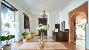 Et hotellobby med reception, lysekrone, potteplanter og et blåt tæppe.
