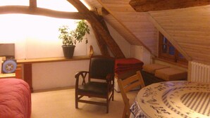 Ein Dachzimmer mit schrägem Dach, ein Holzstuhl, ein Tisch mit gemustertem Tischläufer, eine Topfpflanze und eine rote Decke auf einem Stuhl.