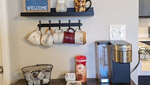 Una encimera de cocina con una cafetera, un cesto de cápsulas de café, un tarro de café y un cartel de bienvenida sobre una estantería con tazas y una planta.