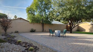Un patio con mesa y sillas, rodeado por un jardín con rocas y cactus, y un edificio al fondo.