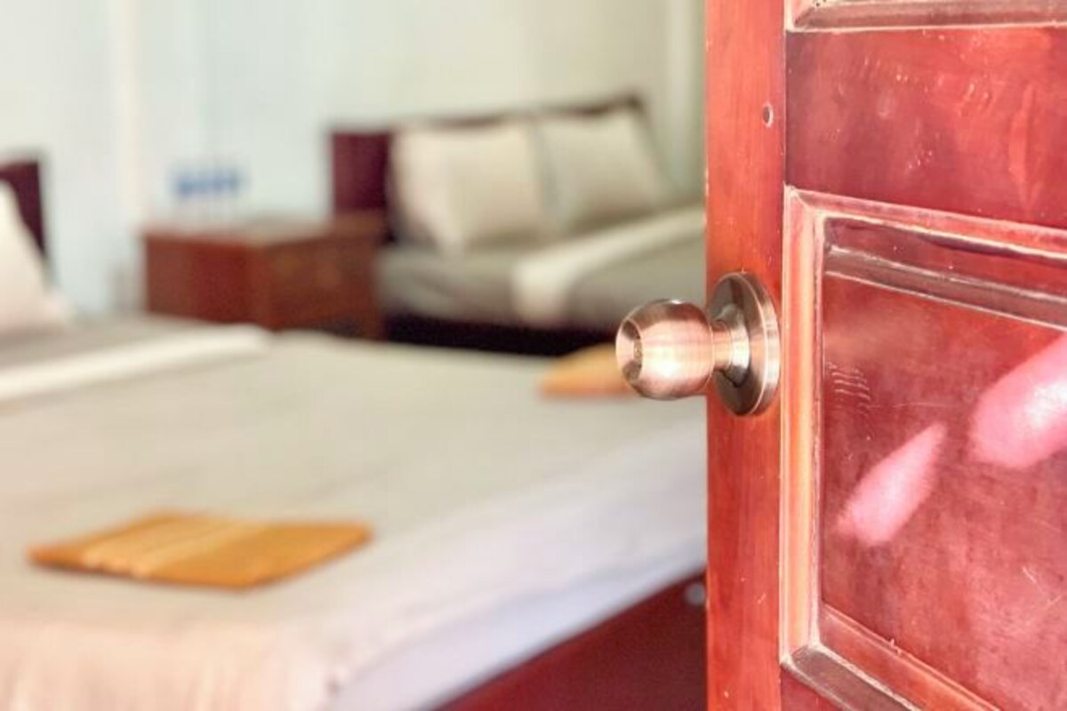 A wooden door with a metallic handle, a bed with a white mattress and a brown blanket, and a nightstand with a lamp.