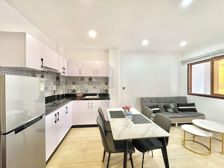 A compact kitchen with a refrigerator, sink, and patterned backsplash. There is a dining table with chairs and a sofa in a living area with a window.
