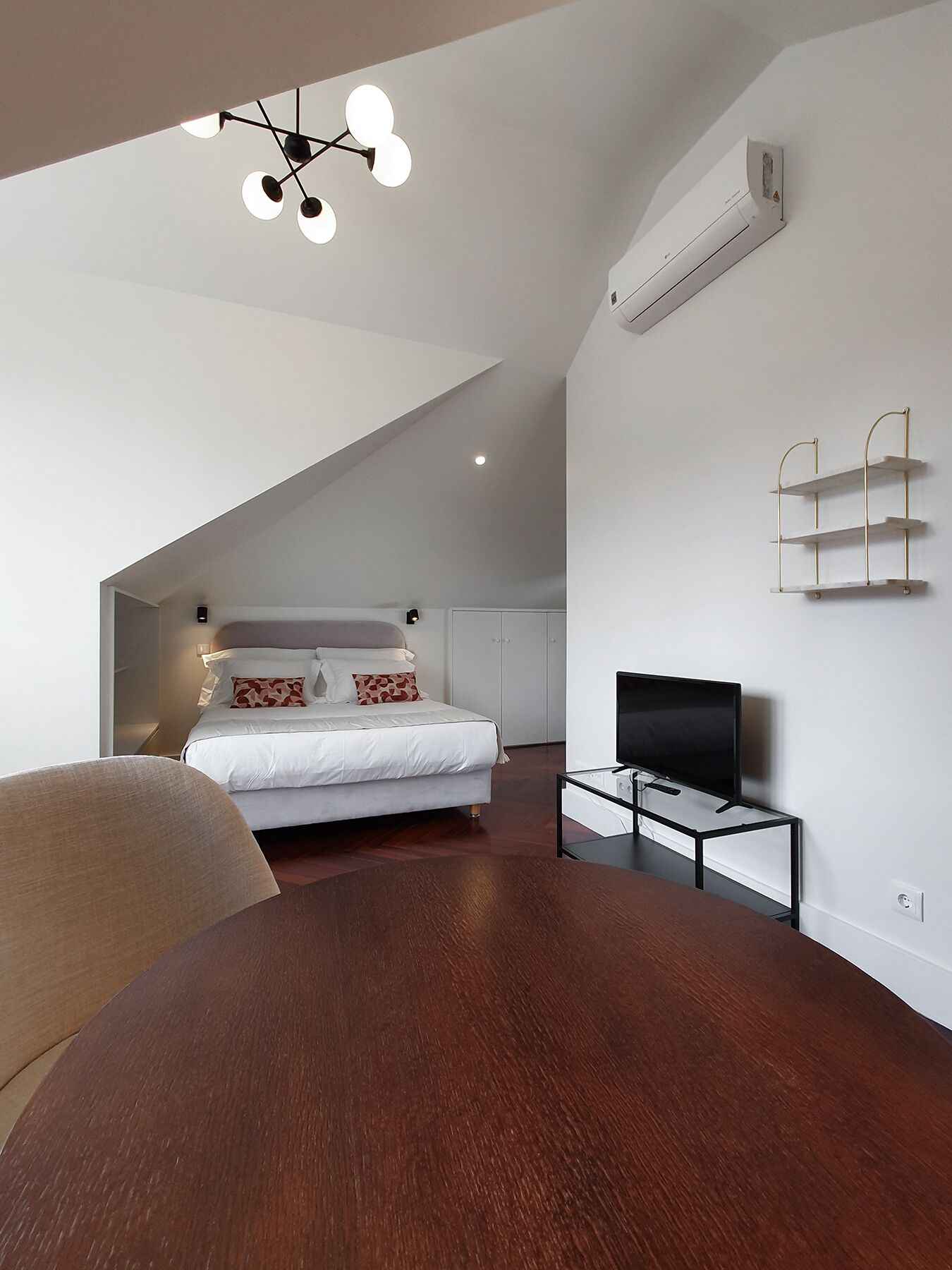 Une chambre moderne avec un plafond en pente, un lit recouvert d’une literie blanche, un téléviseur posé sur un meuble et un luminaire fixé au plafond.