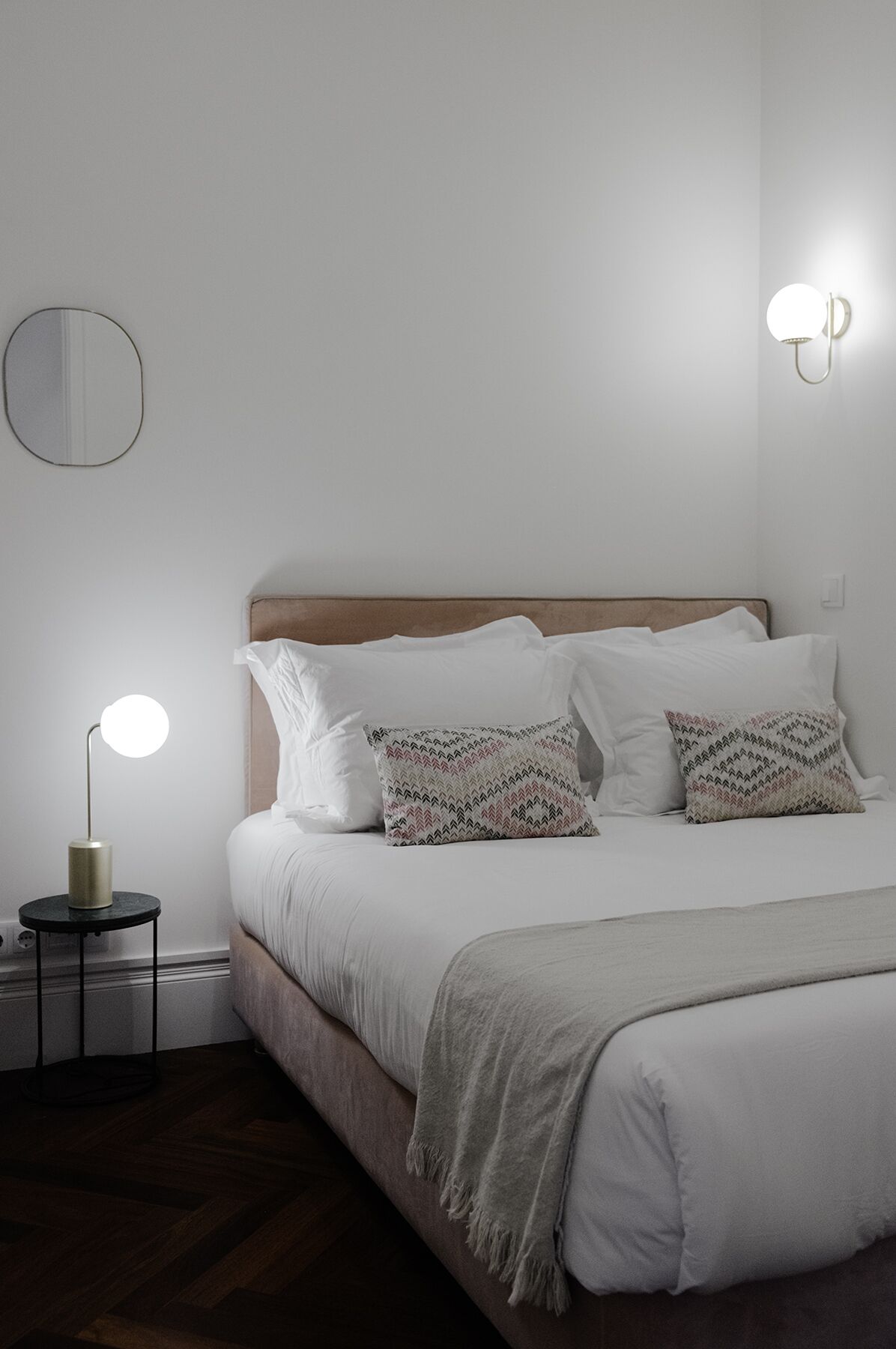 Ein ordentlich gemachtes Bett mit weißer Bettwäsche und gemusterten Kissen, eine Nachttischlampe und ein runder Spiegel an der Wand.